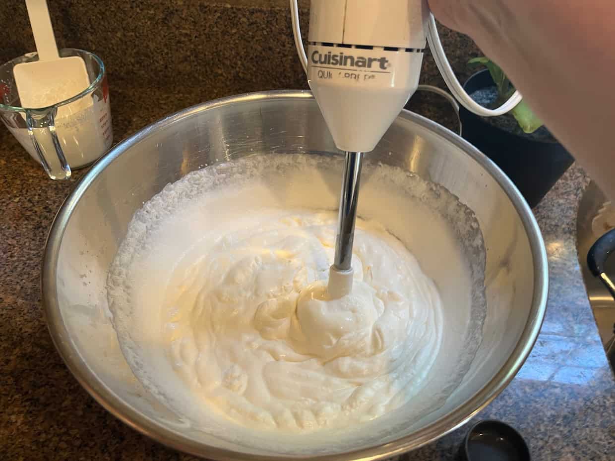 A hand holds a Cuisinart hand mixer blending a thick, white mixture in a large metal bowl on a kitchen counter.