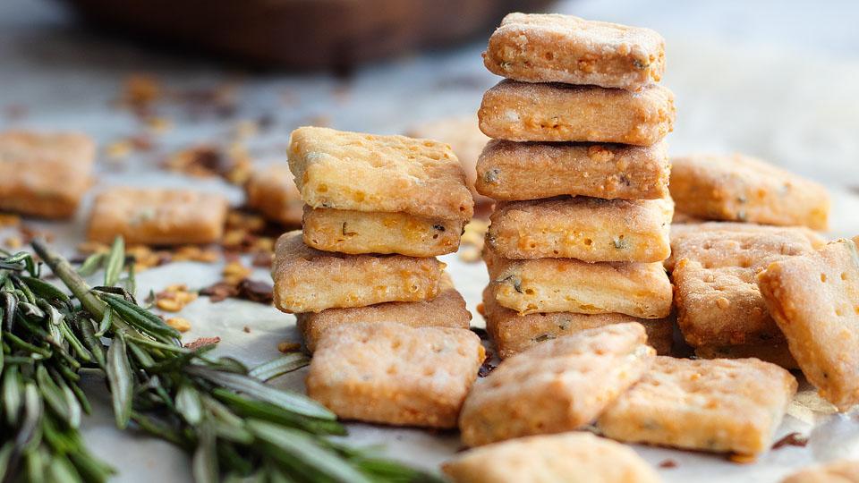 Stacked and scattered square cheese crackers sit on a surface next to sprigs of fresh rosemary.