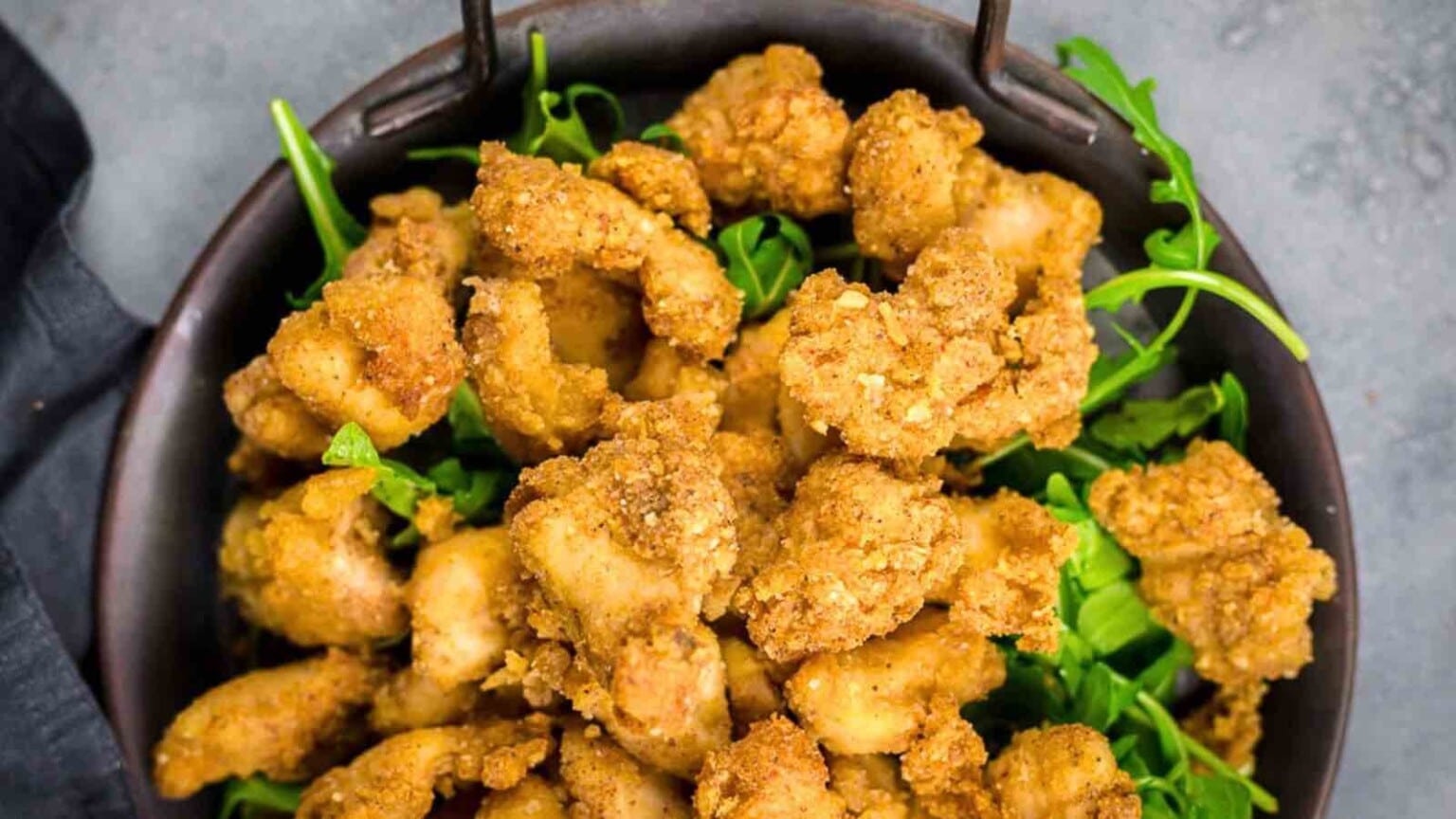 A bowl filled with golden-brown fried chicken pieces served on a bed of fresh green arugula.