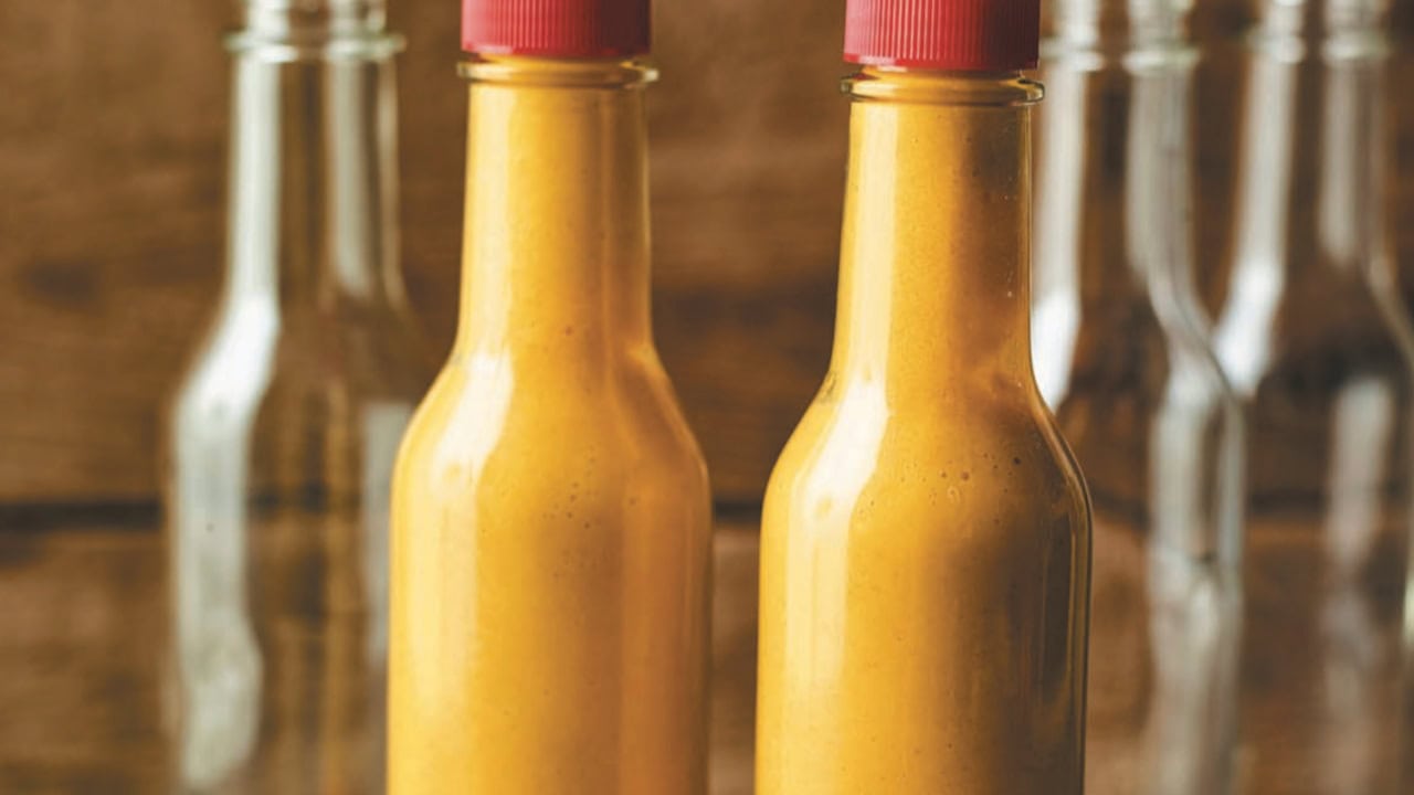 Two filled glass bottles with red caps containing an orange sauce are placed in front of three empty, clear glass bottles.