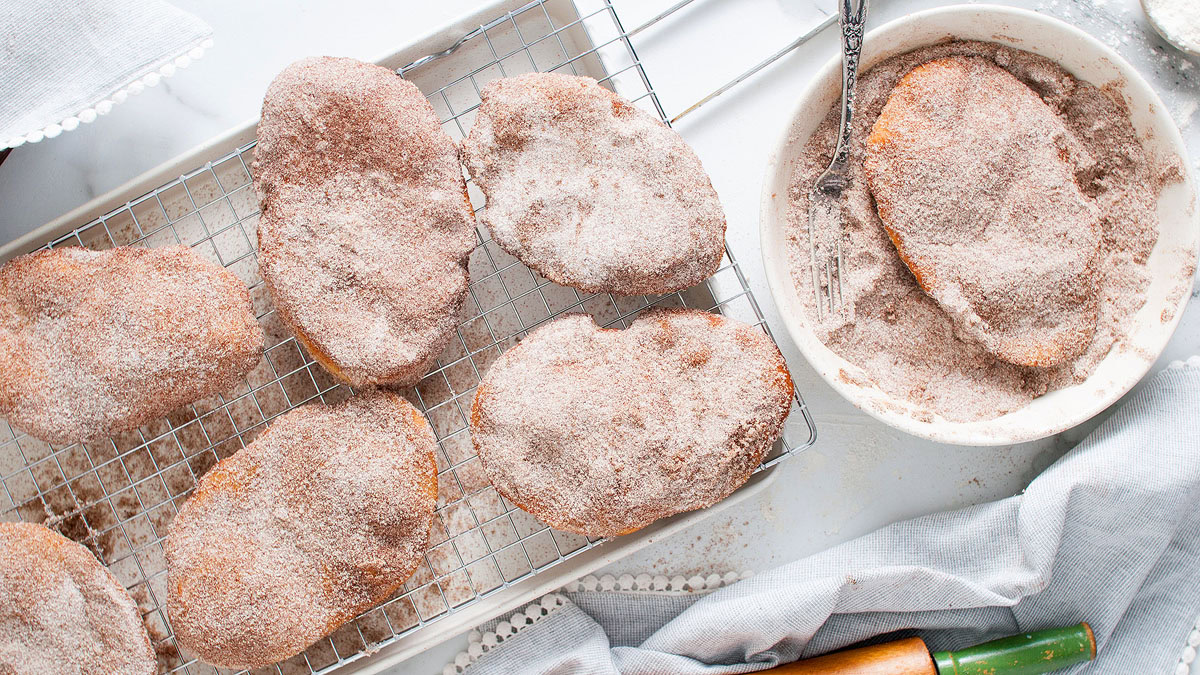 Several pieces of fried dough coated in cinnamon sugar rest on a cooling rack, with one piece being coated in cinnamon sugar in a bowl beside them.