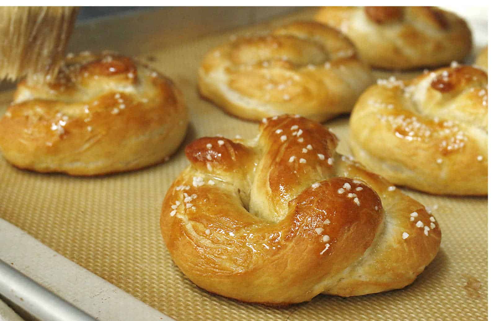 A baking sheet with six golden brown soft pretzels topped with coarse salt, resting on a silicone baking mat.