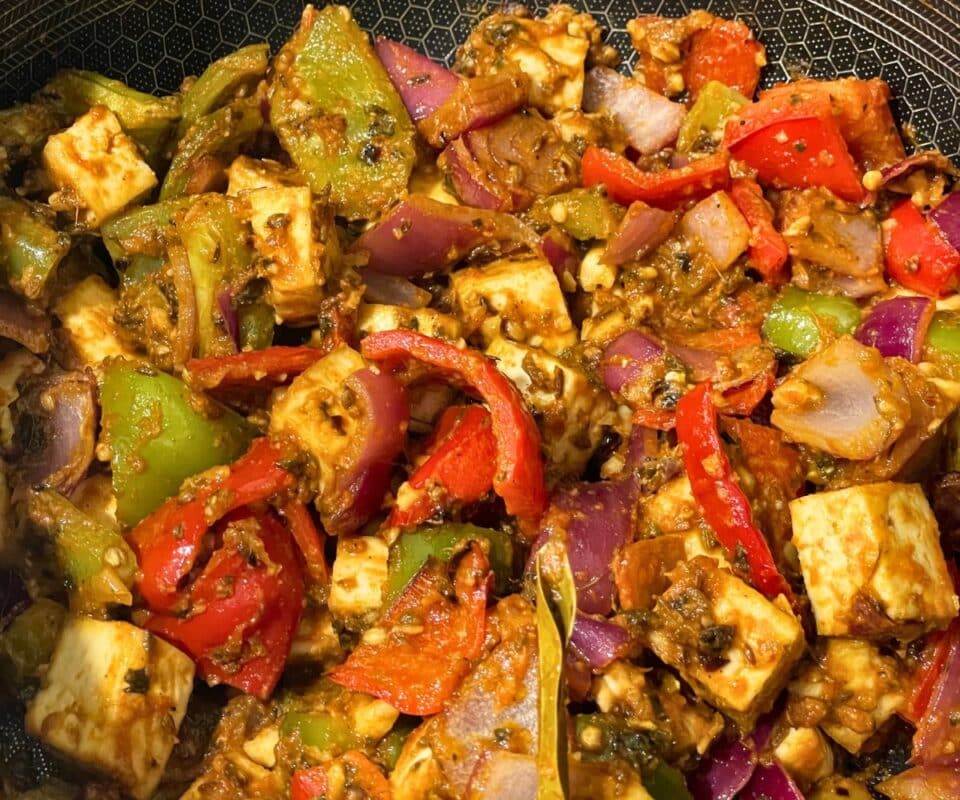 A close-up of a cooked vegetable dish with paneer cubes, red and green bell peppers, and onions in a spiced sauce.