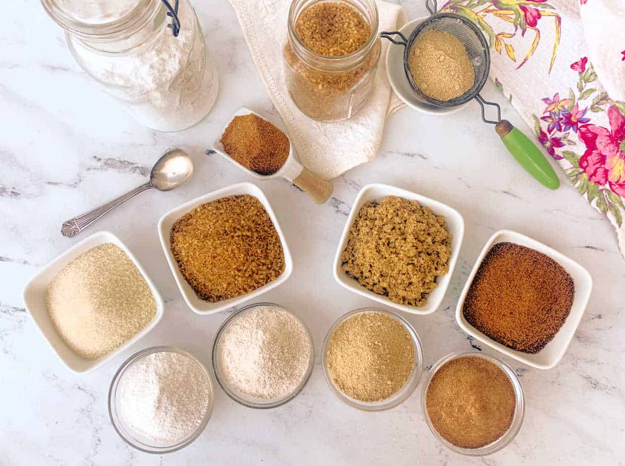 Various types of sugars and flours in jars and bowls are arranged on a white marble surface with a spoon and a floral cloth.