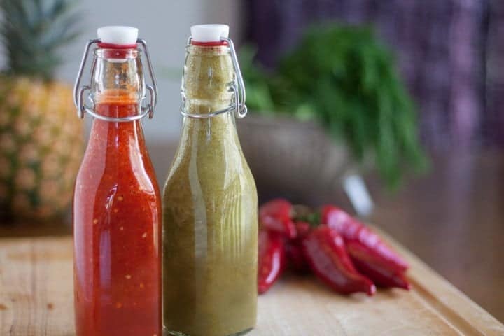 Two glass bottles with clasp lids, one filled with red sauce and the other with green sauce, placed on a wooden surface with peppers and greens in the background.