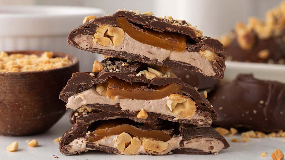 Three chocolate-covered candy pieces are stacked, showing layers of caramel, nougat, whole peanuts, and chocolate coating, with crushed peanuts scattered around.