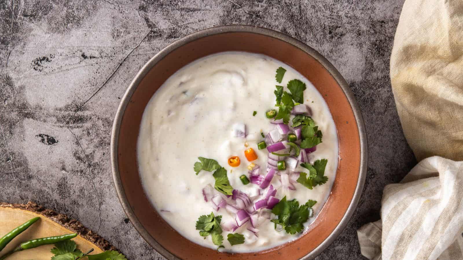 A bowl of raita topped with chopped red onions, green chilies, cilantro, and diced yellow chilies, placed on a gray textured surface.