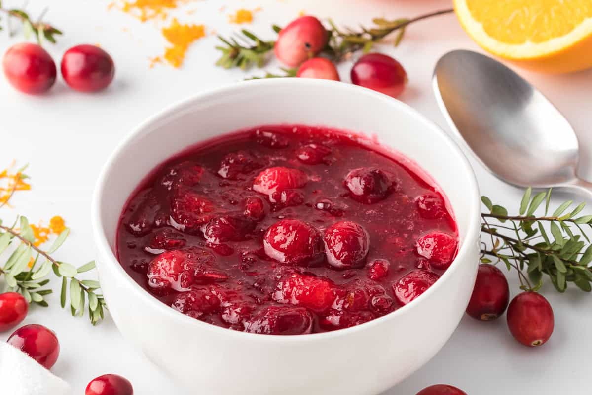 A white bowl filled with cranberry sauce sits on a white surface, surrounded by fresh cranberries, greenery, a spoon, and an orange slice.