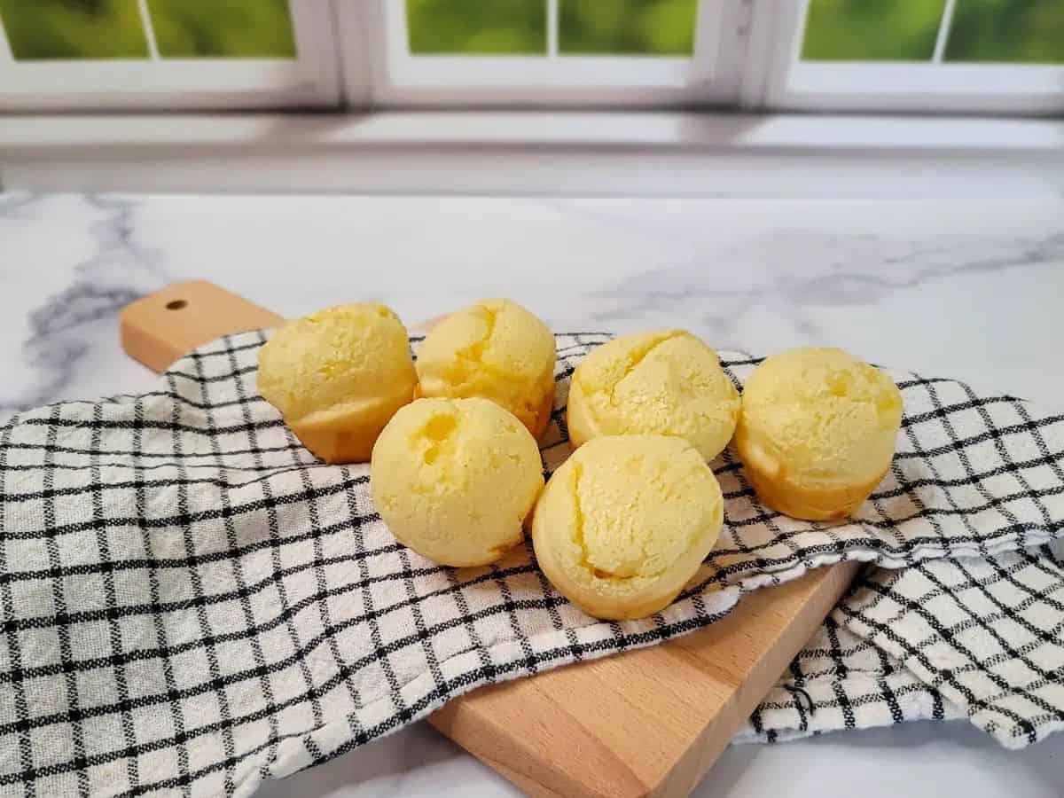 Six small round baked goods are placed on a checkered cloth atop a wooden board, with a window and green scenery in the background.