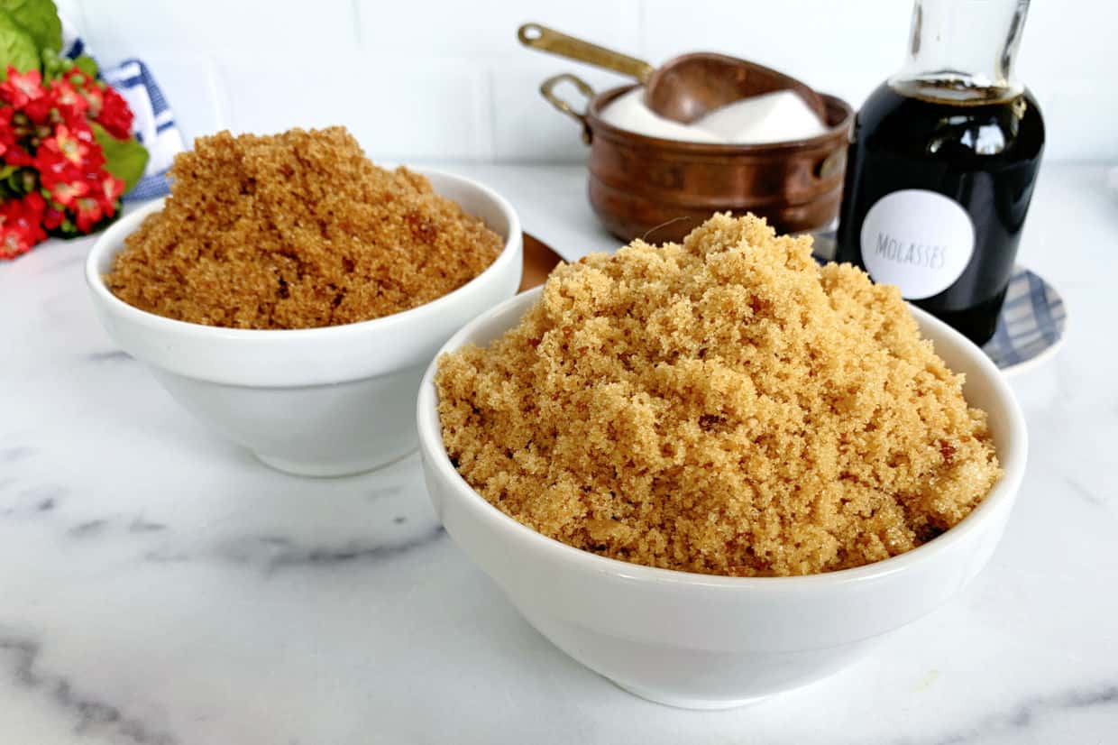 Two white bowls filled with brown sugar are on a marble countertop, with a bottle labeled molasses and a bowl of white sugar in the background.