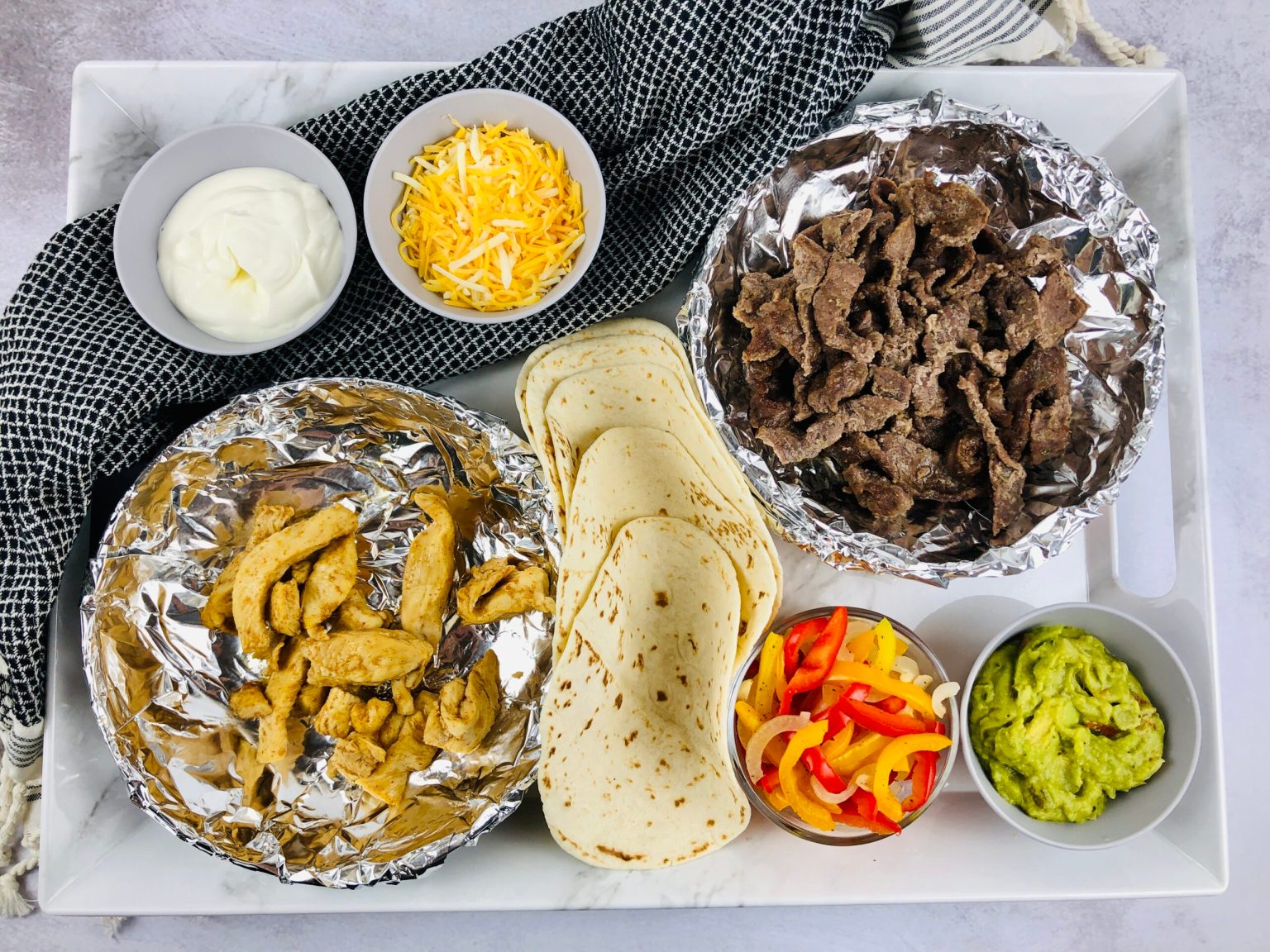 A tray with cooked beef, cooked chicken, flour tortillas, shredded cheese, sour cream, sliced bell peppers, and guacamole in separate bowls and foil containers.