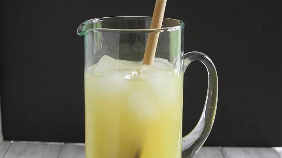 A glass pitcher filled with yellow juice and ice cubes, with a wooden spoon inside, placed on a light-colored surface against a dark background.