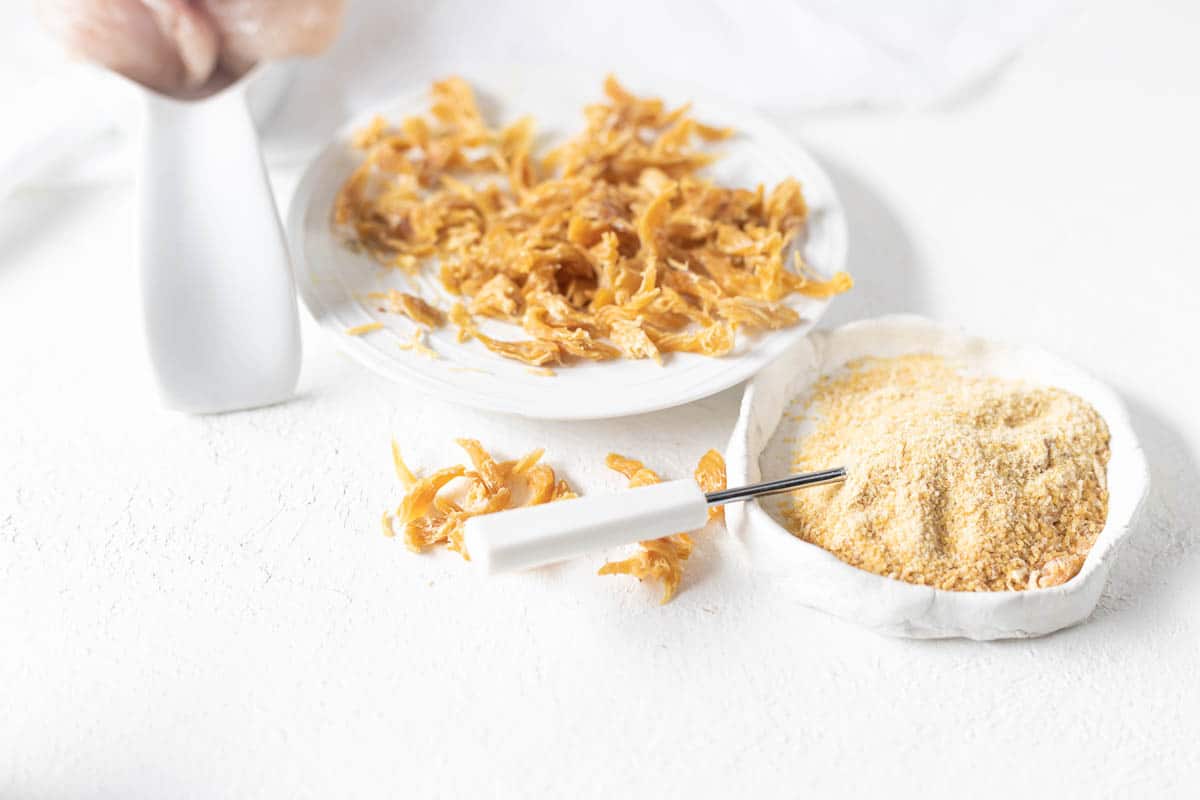 A plate of dried shredded food and a small bowl of powdered crumbs with a grater on a white surface.
