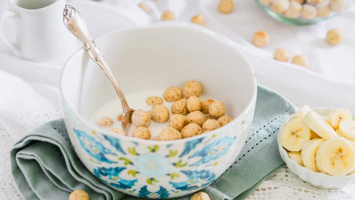 A bowl of milk with round cereal puffs and a spoon, placed on a folded napkin. Sliced bananas are in a small dish nearby.