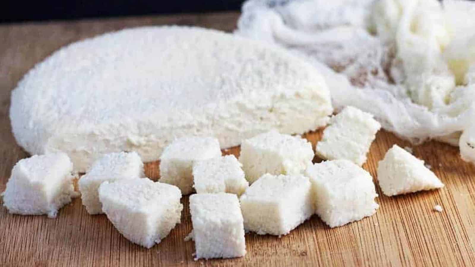 A round block of crumbly white cheese is partially sliced into small cubes, resting on a wooden cutting board next to a piece of cheesecloth.