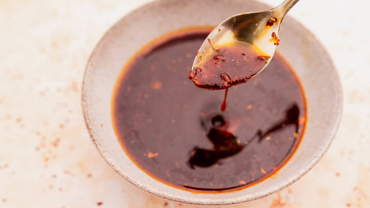 A spoon is held above a bowl filled with dark, oily chili sauce or chili oil, with some of the sauce dripping from the spoon.