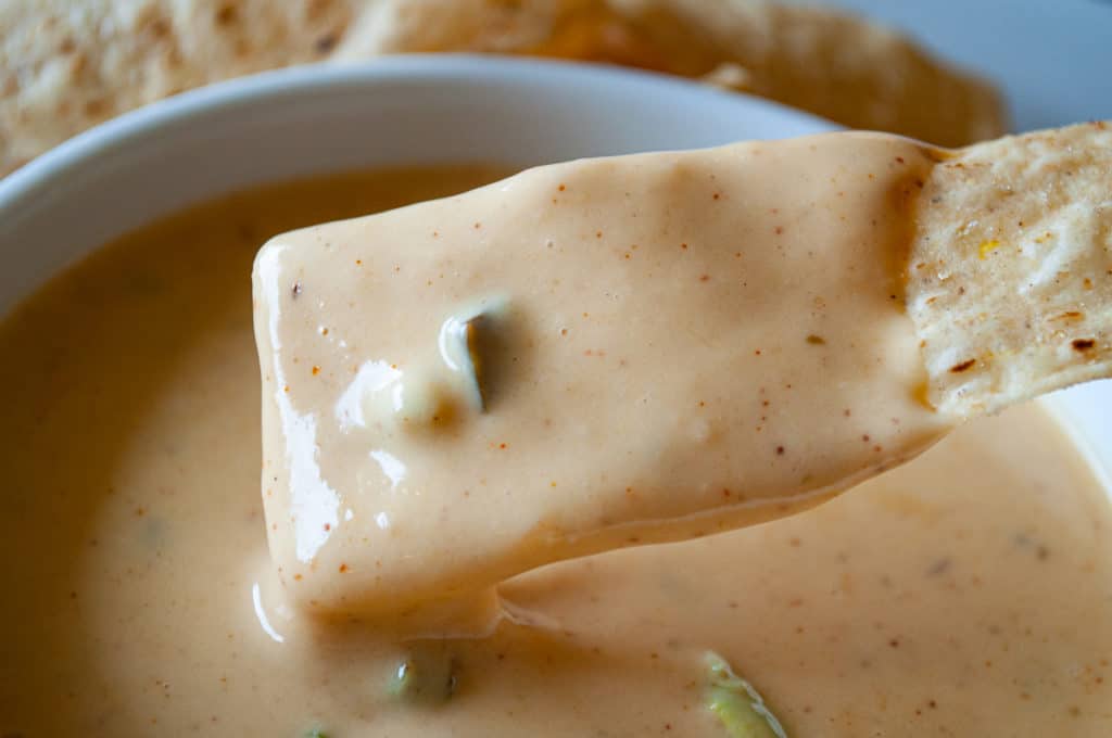 A tortilla chip dipped in creamy, orange queso sauce with visible seasoning and small pieces of green pepper.