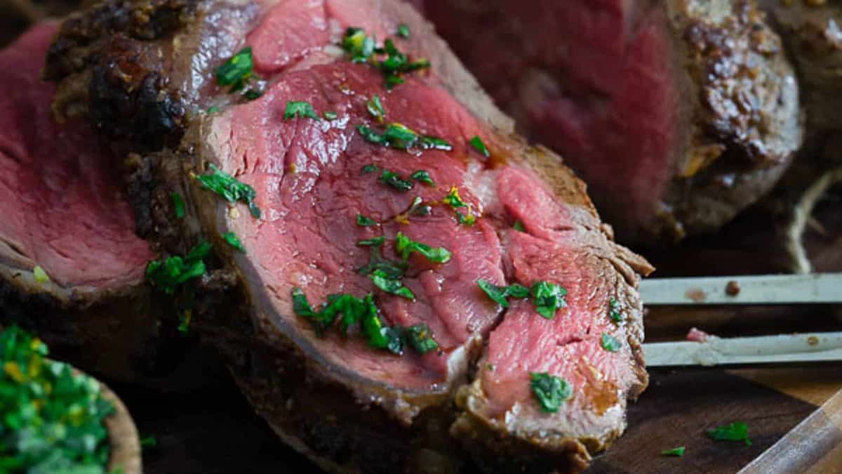 Sliced medium-rare roast beef garnished with chopped herbs, served on a wooden cutting board with carving utensils.