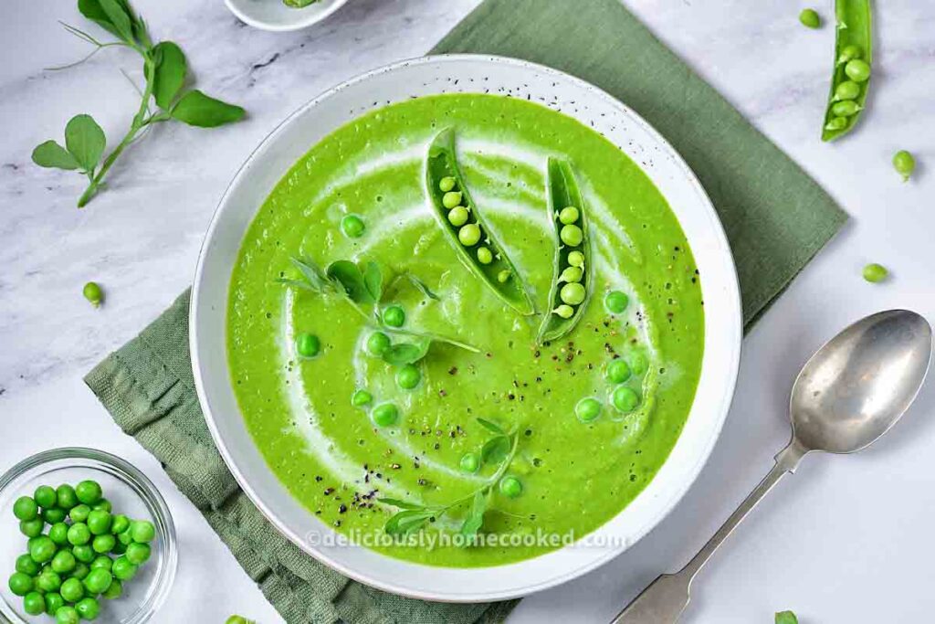 A bowl of green pea soup garnished with pea pods and fresh peas, placed on a green napkin with a spoon and scattered peas beside it.
