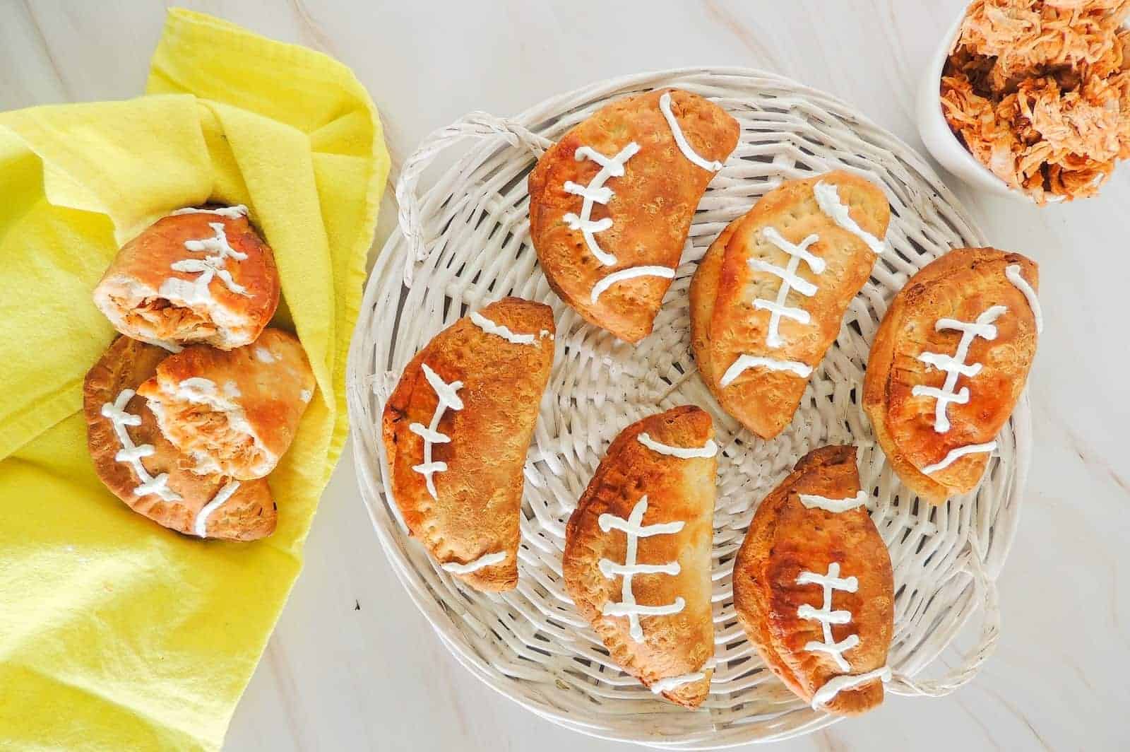 A basket of football-shaped hand pies with white icing laces, next to a yellow napkin holding more pies and a bowl of shredded filling.