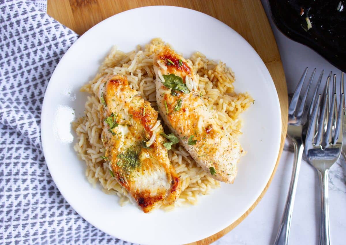 A white plate with two pieces of cooked chicken on a bed of rice, garnished with herbs, next to two forks on a wooden surface.