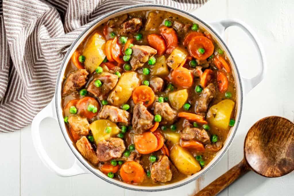 A pot of beef stew with chunks of beef, carrots, potatoes, and peas in a thick broth, next to a wooden spoon and striped towel.