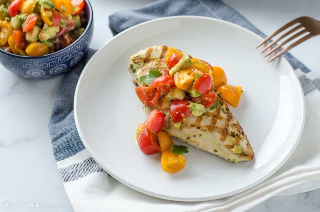 Grilled chicken breast topped with a colorful tomato and avocado salsa on a white plate, with a bowl of extra salsa in the background.