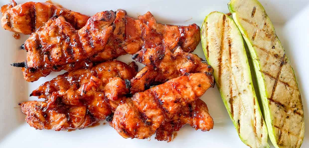 Grilled chicken strips with barbecue sauce served alongside grilled zucchini slices on a white plate.