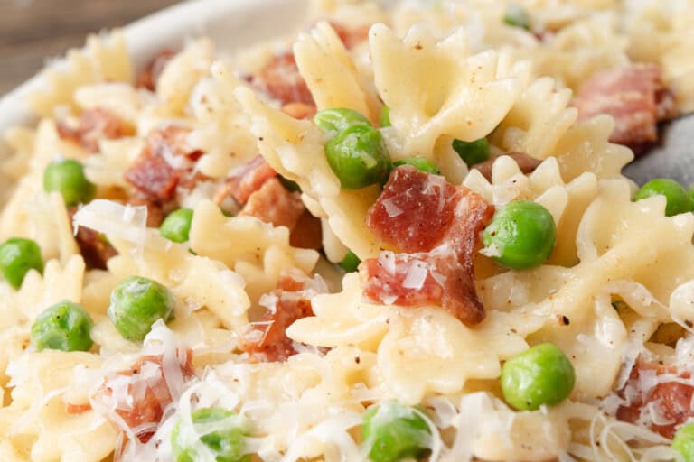Close-up of bow-tie pasta mixed with green peas, crispy bacon pieces, and grated cheese.