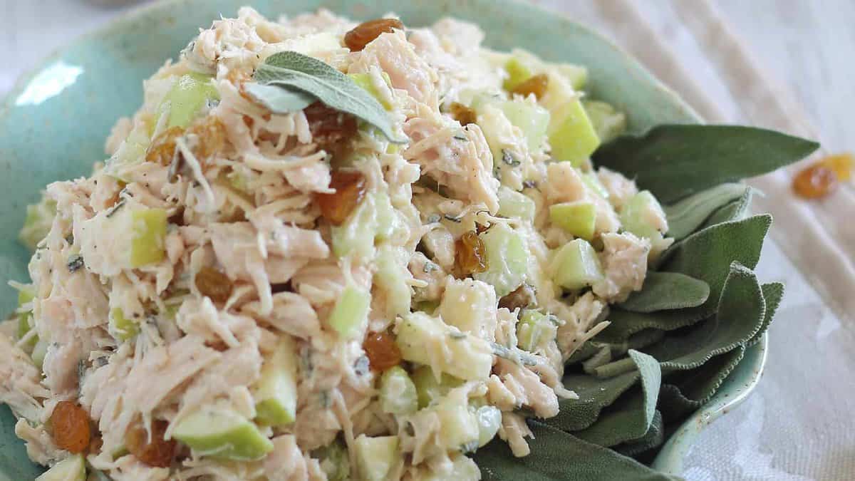 A plate of chicken salad mixed with chopped celery, green herbs, and golden raisins, served on a bed of fresh sage leaves.
