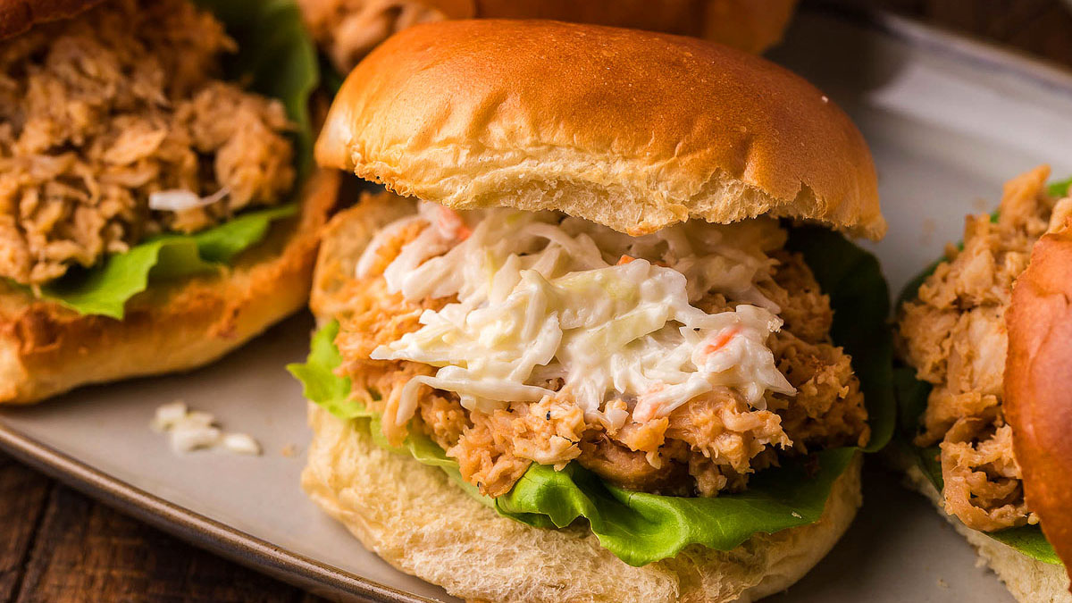 A pulled chicken sandwich with lettuce and coleslaw on a bun, served on a plate.