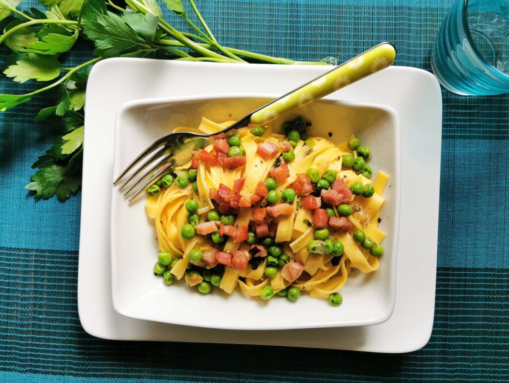 A square white plate with tagliatelle pasta, diced bacon, and green peas, topped with a fork, sits on a blue placemat with parsley and a glass in the background.