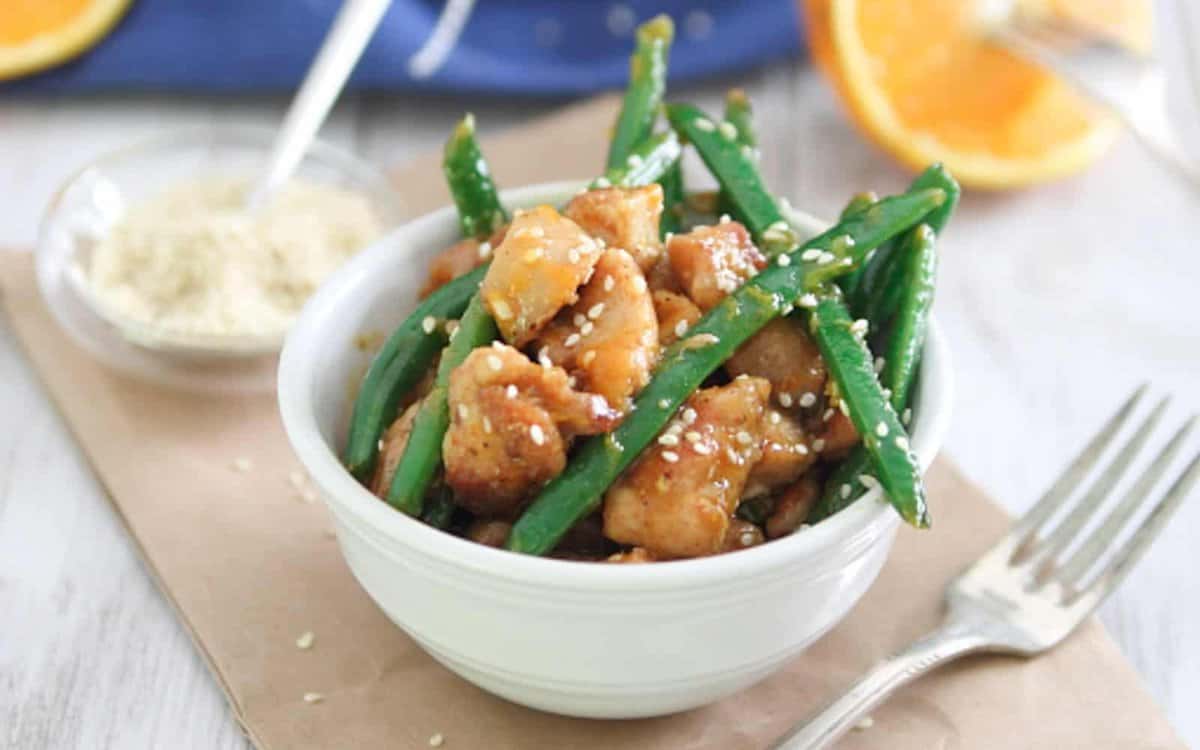 A white bowl filled with cooked chicken pieces and green beans topped with sesame seeds, placed next to a fork and a small dish of white seasoning.