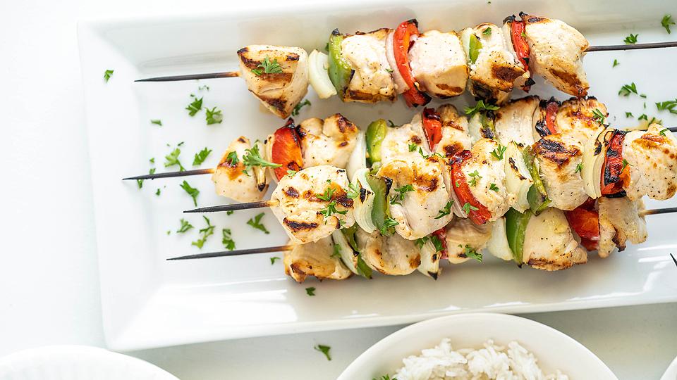 Two grilled chicken kebabs with vegetables on skewers, garnished with chopped herbs, served on a rectangular white plate.