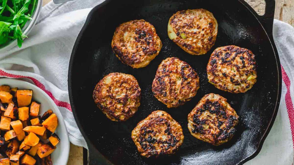 Seven browned turkey patties cooking in a black cast iron skillet, with a plate of roasted sweet potatoes and greens nearby.