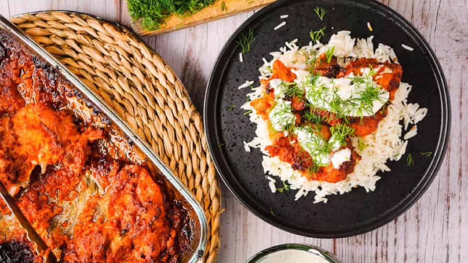 A black plate with white rice topped with a tomato-based baked chicken dish and fresh herbs, next to a baking dish with more of the same chicken.