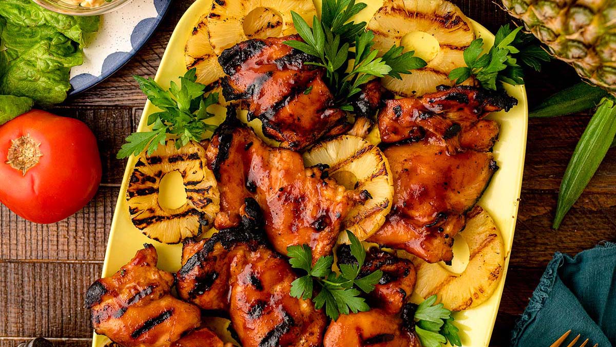 Grilled chicken thighs with barbecue sauce are served on a platter with grilled pineapple rings and garnished with fresh parsley.