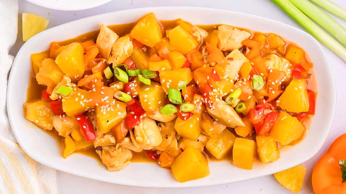 A white oval dish filled with sweet and sour chicken, pineapple chunks, bell peppers, and sauce, garnished with sliced green onions and sesame seeds.