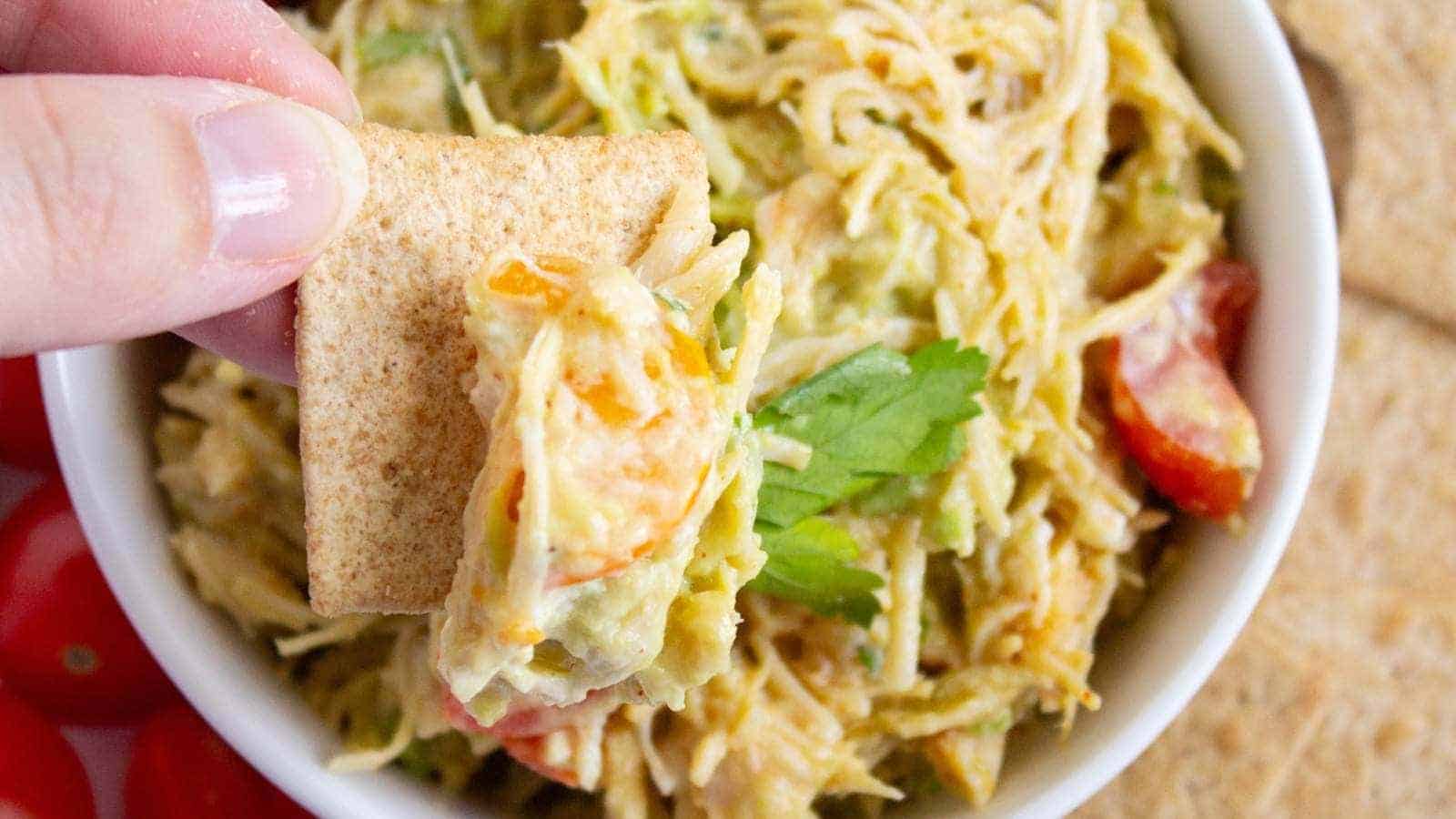 A hand holds a cracker dipped in a bowl of shredded chicken salad with vegetables and a sprig of parsley.