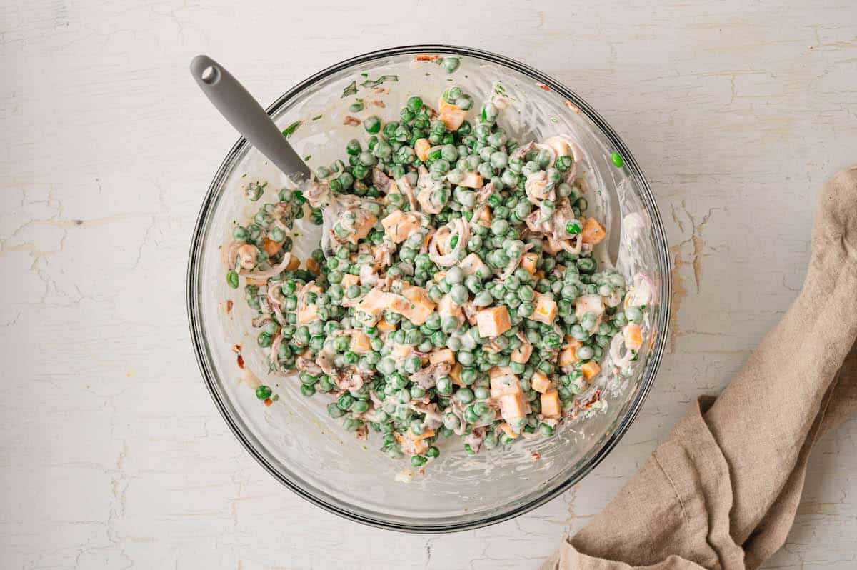 A glass bowl filled with a creamy salad containing peas, diced carrots, cheese cubes, and possibly ham, with a spoon in the bowl and a beige napkin beside it.