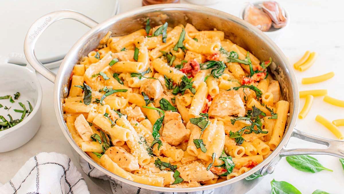Large skillet filled with cooked rigatoni pasta, chicken pieces, spinach, sun-dried tomatoes, and grated cheese, garnished with fresh basil.