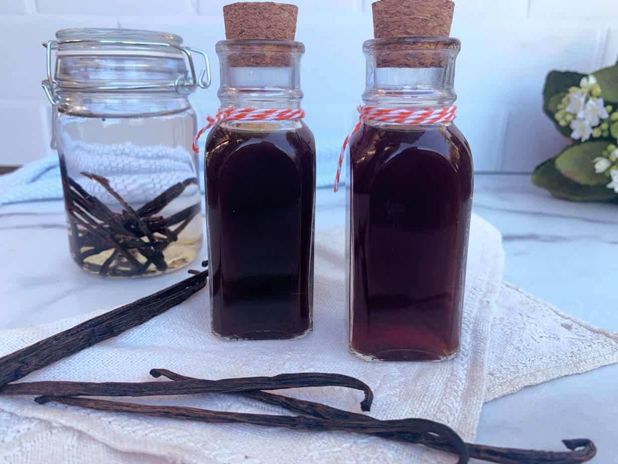 Two small glass bottles filled with dark liquid, sealed with corks and twine, next to vanilla beans and a jar with vanilla pods soaking in liquid.