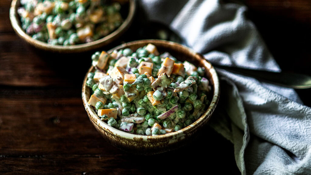 A bowl of pea salad with chopped cheese, red onions, and creamy dressing on a dark wooden table beside a folded cloth napkin.