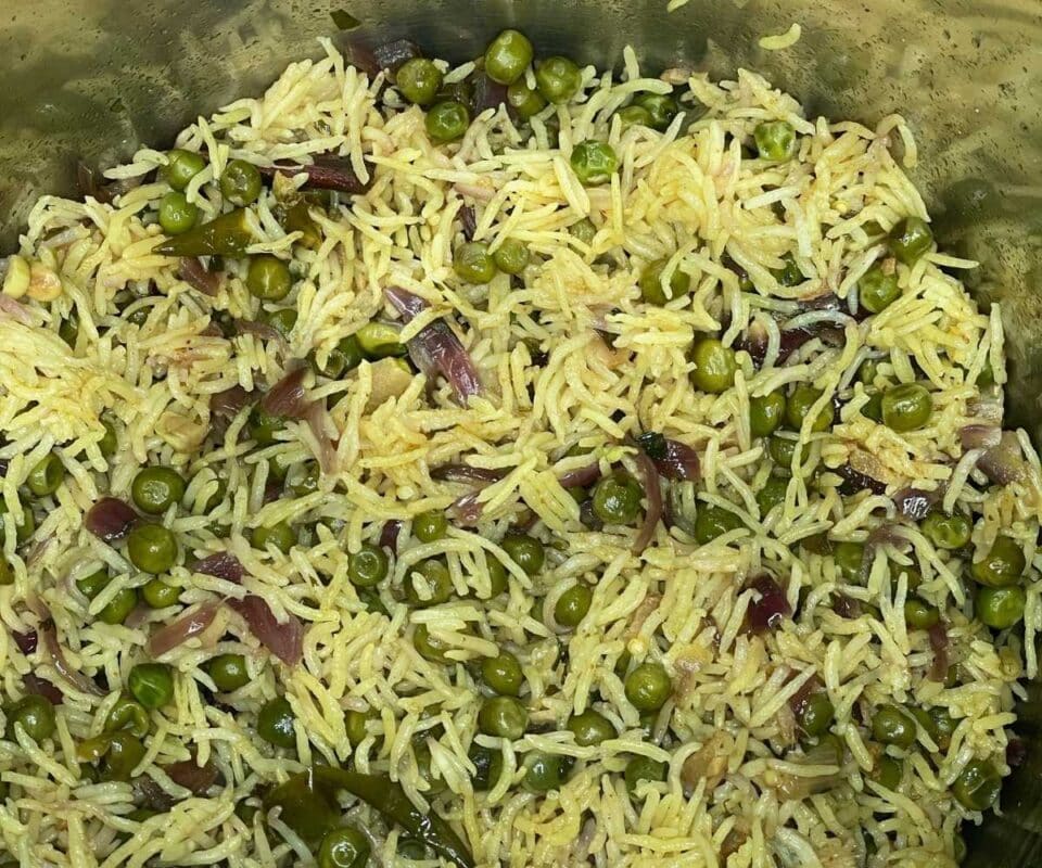 Cooked rice mixed with green peas, sliced onions, and spices in a large pot.