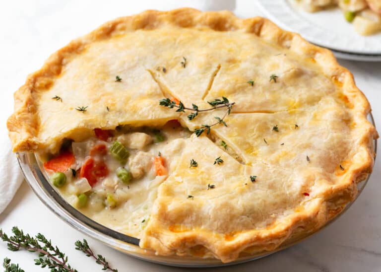 A baked chicken pot pie with a golden crust, partially sliced to reveal a filling of chicken, peas, carrots, and creamy sauce, garnished with fresh thyme.