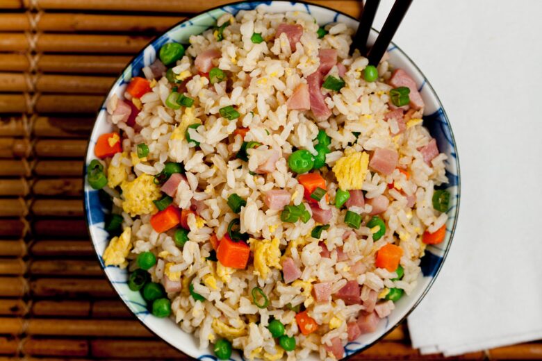 A bowl of fried rice with diced ham, scrambled eggs, peas, carrots, and green onions, served with chopsticks on a bamboo mat.