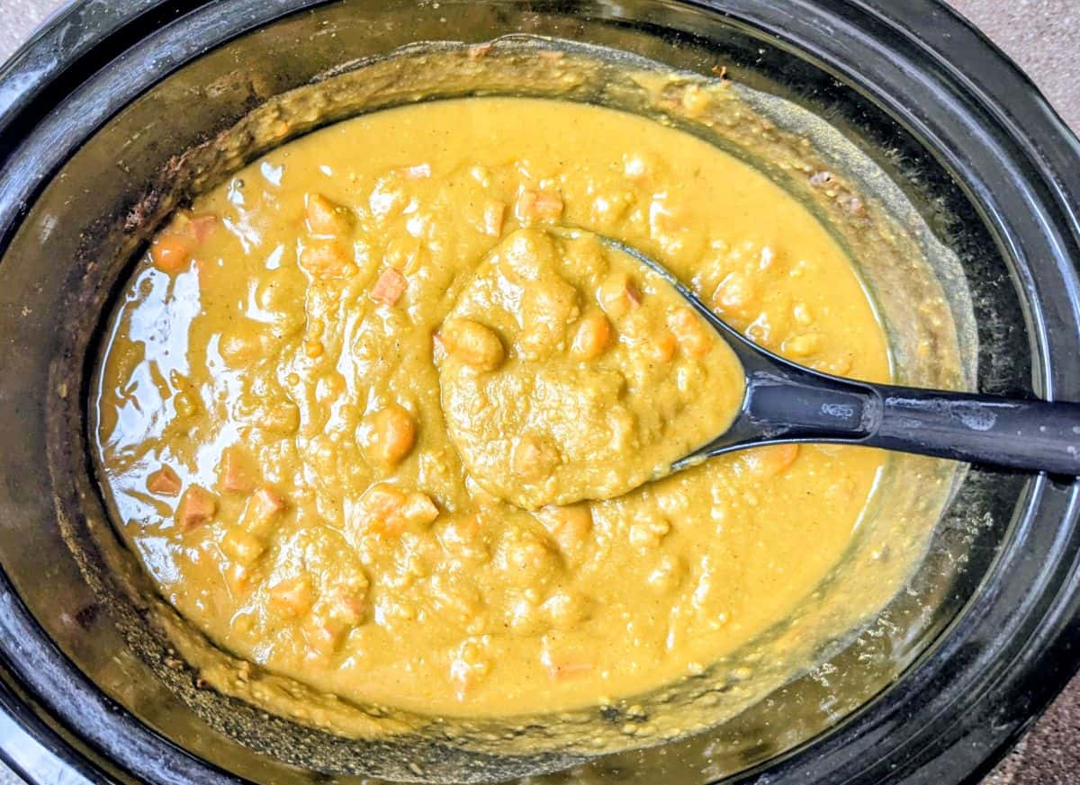 A slow cooker filled with thick yellow split pea soup, with visible chunks and a black serving spoon resting inside.