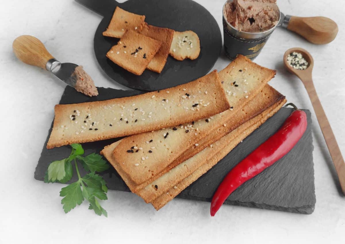 Rectangular sesame crackers on a slate board with a red chili, parsley, pรขtรฉ, and serving utensils.
