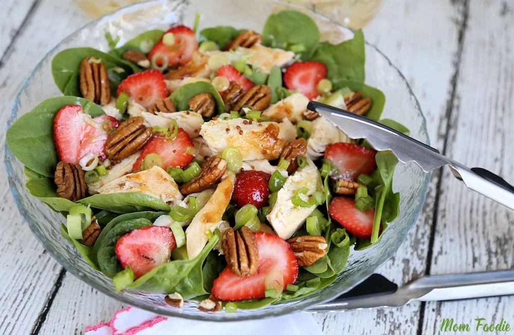A glass bowl with spinach salad topped with grilled chicken, strawberries, pecans, and green onions. Metal tongs rest on the edge of the bowl.