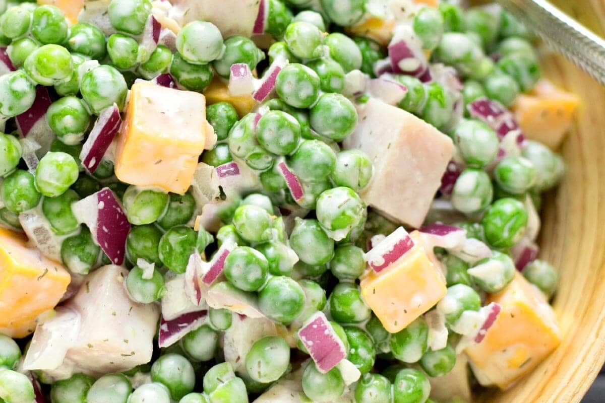 Close-up of a salad with green peas, diced ham, cheddar cheese cubes, chopped red onion, and a creamy dressing.